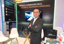 AMD lleva la Infraestructura de IA más allá de los centros de datos Fotografía en plano medio que muestra a Hiram Monroy, Sr Manager de AMD, posando de frente con los brazos cruzados en un stand corporativo. El ejecutivo viste un traje de color gris Oxford, camisa blanca y anteojos de armazón oscuro. Detrás de él, un muro publicitario negro con bordes iluminados por luces LED verdes exhibe la inscripción "IMPULSA LA INNOVACIÓN CON IA, ACELERA EL CRECIMIENTO EMPRESARIAL", junto a una pantalla digital central. A los lados se aprecian sillas de diseño blancas, una mesa de cristal y artículos promocionales de la marca que incluyen una bolsa negra con el lema "together we advance_AI", una playera de fútbol y un balón deportivo.