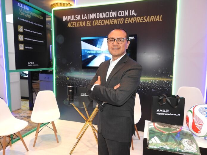 Fotografía en plano medio que muestra a Hiram Monroy, Sr Manager de AMD, posando de frente con los brazos cruzados en un stand corporativo. El ejecutivo viste un traje de color gris Oxford, camisa blanca y anteojos de armazón oscuro. Detrás de él, un muro publicitario negro con bordes iluminados por luces LED verdes exhibe la inscripción 