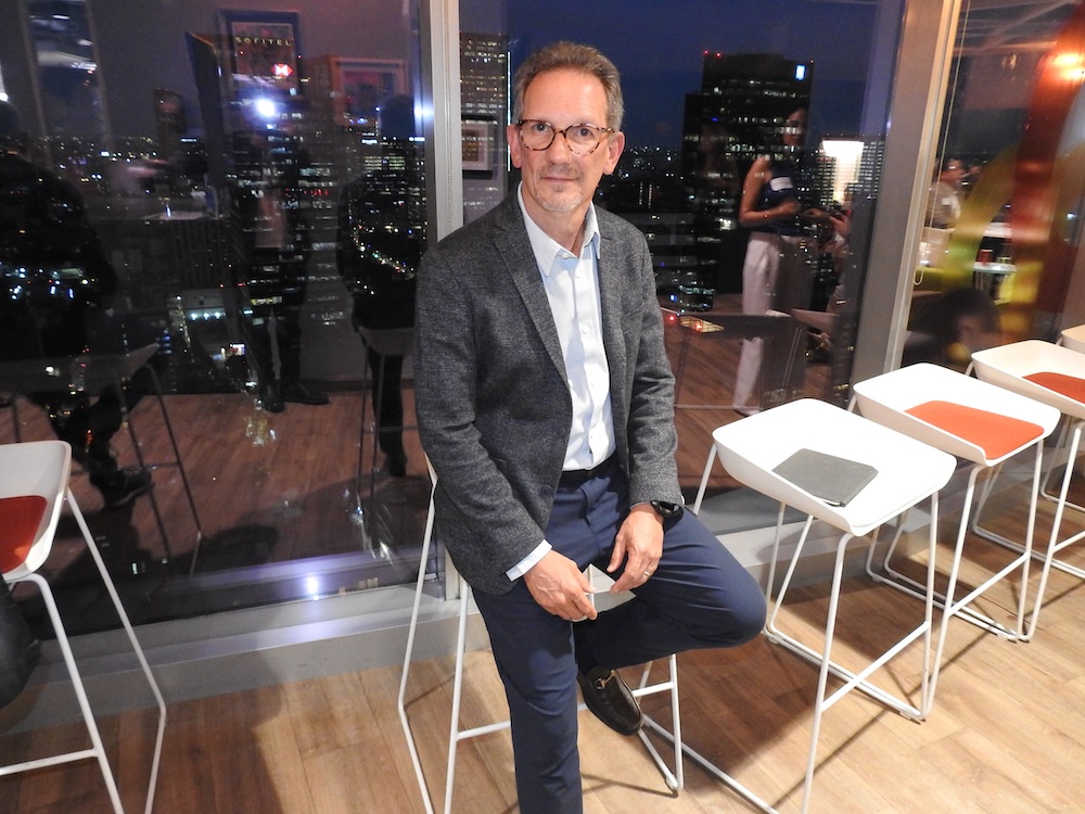 Fotografía en plano medio de Javier Cordero, VP y General Manager North of Latin America en Red Hat. El ejecutivo, con lentes de armazón oscuro, viste un saco gris texturizado sobre una camisa a rayas claras y un pantalón azul marino. Se encuentra sentado sobre un taburete alto blanco en un espacio interior con piso de madera, teniendo detrás un gran ventanal que muestra una vista nocturna difuminada de los rascacielos iluminados de la Ciudad de México.