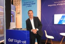 Digit-all consolida su propuesta de ciberseguridad para empresas con nube e inteligencia artificial Fotografía en plano medio largo que muestra a Jesús González, responsable del área de producto y preventa en Digit-all, posando de frente y ligeramente inclinado al apoyarse en el mostrador azul de su stand corporativo. El directivo viste un saco negro casual sobre una camisa blanca de cuello abierto y pantalón oscuro, manteniendo las manos entrelazadas al frente. Detrás de él, la columna del stand exhibe el letrero "Expertos en ciberseguridad y nube" sobre un fondo azul claro, flanqueado por una pantalla digital y una silla blanca minimalista.