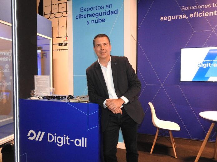 Jesús González detalla el modelo de acompañamiento técnico para la mitigación de riesgos digitales en corporativos Fotografía en plano medio largo que muestra a Jesús González, responsable del área de producto y preventa en Digit-all, posando de frente y ligeramente inclinado al apoyarse en el mostrador azul de su stand corporativo. El directivo viste un saco negro casual sobre una camisa blanca de cuello abierto y pantalón oscuro, manteniendo las manos entrelazadas al frente. Detrás de él, la columna del stand exhibe el letrero