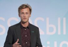 HPE revela el elefante blanco en la sala de servidores: la huella energética de la computación tradicional Fotografía en plano medio que muestra a Jostein Birkeland, HPE Principal Technologist en Transformación Sostenible, hablando de frente durante una conferencia de infraestructura digital. El especialista posee cabello rubio corto y barba ligera, y viste un saco sastre gris oscuro sobre una camisa de vestir negra con pequeños lunares blancos en patrón regular. En la solapa izquierda de su saco resalta un discreto pin rectangular verde con el logotipo oficial de Hewlett Packard Enterprise. Mantiene su mano derecha levantada realizando un gesto con los dedos abiertos como apoyo a su discurso, mientras que al fondo se observa de forma difuminada un panel con letras blancas sobre una superficie azul claro uniforme.