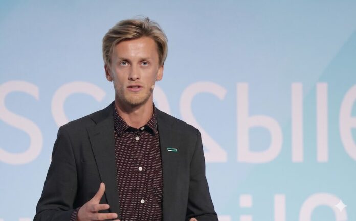 Jostein Birkeland analiza el verdadero impacto de la computación convencional en la huella energética global de TI Fotografía en plano medio que muestra a Jostein Birkeland, HPE Principal Technologist en Transformación Sostenible, hablando de frente durante una conferencia de infraestructura digital. El especialista posee cabello rubio corto y barba ligera, y viste un saco sastre gris oscuro sobre una camisa de vestir negra con pequeños lunares blancos en patrón regular. En la solapa izquierda de su saco resalta un discreto pin rectangular verde con el logotipo oficial de Hewlett Packard Enterprise. Mantiene su mano derecha levantada realizando un gesto con los dedos abiertos como apoyo a su discurso, mientras que al fondo se observa de forma difuminada un panel con letras blancas sobre una superficie azul claro uniforme.