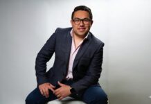 Stulz destaca el papel de la climatización de precisión en el crecimiento de los data centers en Perú Fotografía de estudio en plano medio de Josue Misael González, Sales Executive LATAM en Stulz, posando sentado e inclinado hacia adelante sobre un fondo blanco grisáceo claro uniforme. Viste un saco de vestir texturizado azul marino, una camisa de color rosa pálido con el cuello abierto y pantalones de mezclilla oscuros. El ejecutivo utiliza anteojos de armazón negro, lleva un reloj inteligente negro en su muñeca izquierda y sonríe directamente hacia la cámara.
