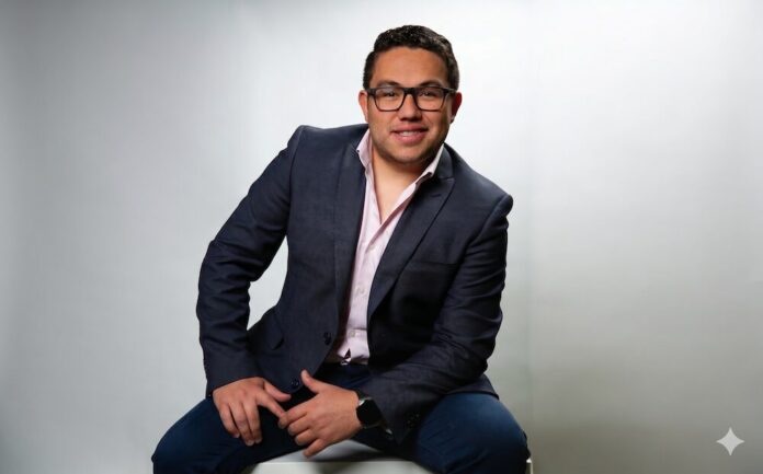 Fotografía de estudio en plano medio de Josue Misael González, Sales Executive LATAM en Stulz, posando sentado e inclinado hacia adelante sobre un fondo blanco grisáceo claro uniforme. Viste un saco de vestir texturizado azul marino, una camisa de color rosa pálido con el cuello abierto y pantalones de mezclilla oscuros. El ejecutivo utiliza anteojos de armazón negro, lleva un reloj inteligente negro en su muñeca izquierda y sonríe directamente hacia la cámara.