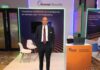 Servicios de ciberseguridad de Akamai abren nuevas oportunidades para telcos Fotografía en plano medio que muestra a un directivo posando de frente con las manos en los bolsillos de su pantalón dentro de un stand corporativo. El ejecutivo viste un traje sastre oscuro, camisa blanca de vestir y una corbata roja con patrones geométricos sutiles. A sus espaldas resalta un muro publicitario con fondo de ondas naranjas y tipografía blanca bajo el encabezado "Akamai Security", flanqueado a la derecha por mesas de atención y un mostrador naranja que exhibe una computadora portátil abierta.