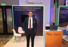 Servicios de ciberseguridad de Akamai abren nuevas oportunidades para telcos Fotografía en plano medio que muestra a un directivo posando de frente con las manos en los bolsillos de su pantalón dentro de un stand corporativo. El ejecutivo viste un traje sastre oscuro, camisa blanca de vestir y una corbata roja con patrones geométricos sutiles. A sus espaldas resalta un muro publicitario con fondo de ondas naranjas y tipografía blanca bajo el encabezado "Akamai Security", flanqueado a la derecha por mesas de atención y un mostrador naranja que exhibe una computadora portátil abierta.