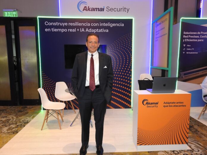Juan José Quintanar detalla la evolución arquitectónica de Akamai hacia los servicios cloud y la seguridad avanzada Fotografía en plano medio que muestra a un directivo posando de frente con las manos en los bolsillos de su pantalón dentro de un stand corporativo. El ejecutivo viste un traje sastre oscuro, camisa blanca de vestir y una corbata roja con patrones geométricos sutiles. A sus espaldas resalta un muro publicitario con fondo de ondas naranjas y tipografía blanca bajo el encabezado