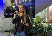 Ingram Micro y Microsoft presentan la estrategia Edge to Cloud para una infraestructura inteligente Fotografía en plano medio que captura a Juanita Gómez, Sr. Windows Commercial Category Lead Americas para Microsoft, hablando de frente con un micrófono inalámbrico en la mano derecha durante una conferencia tecnológica. La ejecutiva, de tez clara y cabello largo castaño claro, viste una blusa negra con saco oscuro casual de corte asimétrico y pantalón de mezclilla azul. En su mano izquierda sostiene un pasador de diapositivas, y en su muñeca lleva un reloj inteligente y pulseras. Detrás de ella resalta una gran pantalla digital que despliega la diapositiva "The Hybrid..." bajo el logotipo de Windows 11 y Copilot+ PC, mientras a la derecha se aprecia vegetación ornamental y ventanales con iluminación natural.