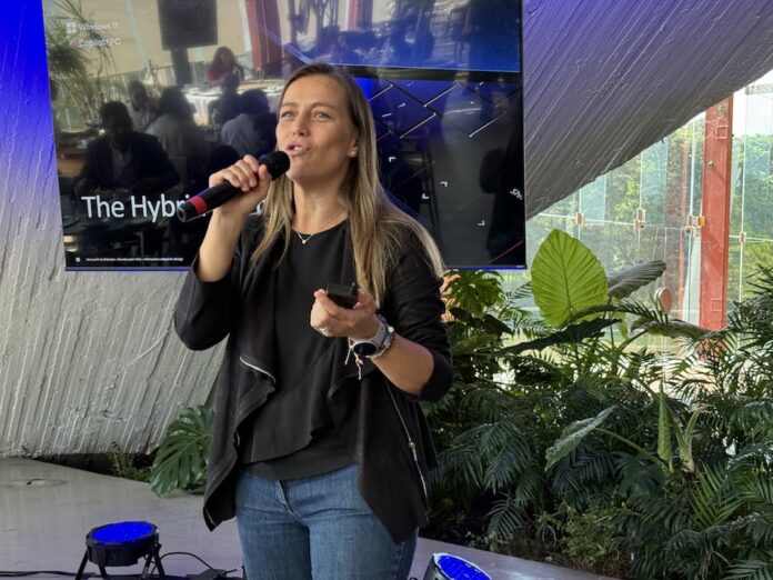 Fotografía en plano medio que captura a Juanita Gómez, Sr. Windows Commercial Category Lead Americas para Microsoft, hablando de frente con un micrófono inalámbrico en la mano derecha durante una conferencia tecnológica. La ejecutiva, de tez clara y cabello largo castaño claro, viste una blusa negra con saco oscuro casual de corte asimétrico y pantalón de mezclilla azul. En su mano izquierda sostiene un pasador de diapositivas, y en su muñeca lleva un reloj inteligente y pulseras. Detrás de ella resalta una gran pantalla digital que despliega la diapositiva 