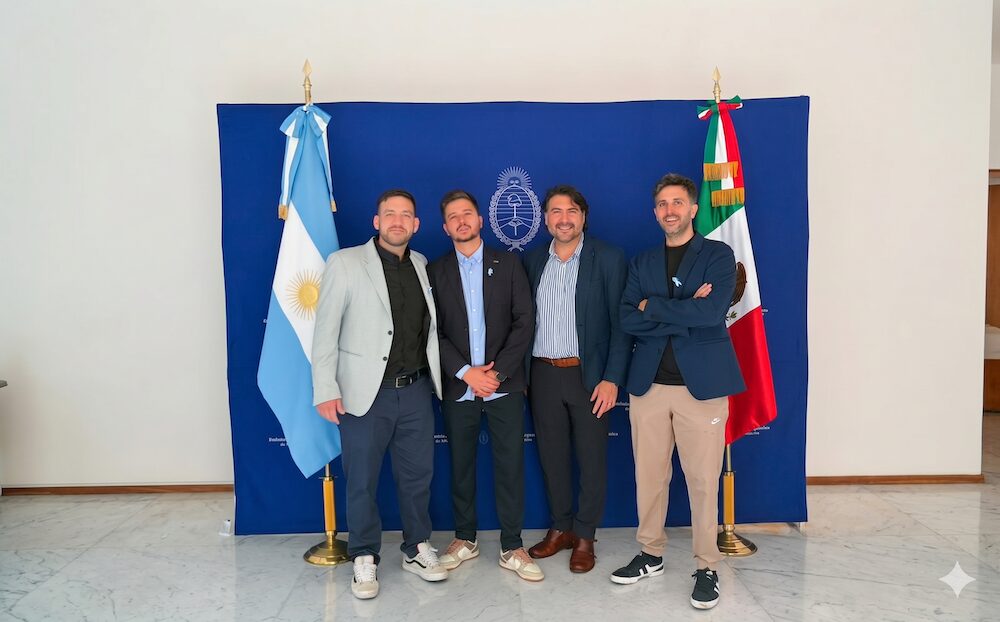 Inteligencia artificial empresarial: Fotografía de plano entero que muestra a cuatro directivos masculinos posando alineados de pie ante un gran fondo de tela azul rey que exhibe en el centro el escudo oficial de la República Argentina en color blanco. En los extremos se sitúan pedestales con banderas: a la izquierda la bandera nacional argentina y a la derecha la bandera nacional de México. Los profesionales visten ropa casual de negocios y portan pequeños lazos celestes prendidos en sus sacos.
