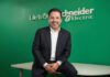 De la digitalización a la infraestructura crítica: el gran reto de la transformación Fotografía de estudio en plano medio que muestra a José Alberto Llavot, directivo de Schneider Electric, posando sentado e inclinado hacia adelante sobre el borde de un escritorio blanco. El ejecutivo, de cabello oscuro y barba ligera, viste un saco de traje negro, camisa blanca de cuello abierto y mantiene las manos entrelazadas al frente con una sonrisa clara hacia la cámara. Detrás de él, un muro liso de color verde brillante exhibe el logotipo institucional de "Schneider Electric" junto a la frase representativa de la marca "Life Is On" en letras blancas en relieve.