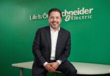 De la digitalización a la infraestructura crítica: el gran reto de la transformación Fotografía de estudio en plano medio que muestra a José Alberto Llavot, directivo de Schneider Electric, posando sentado e inclinado hacia adelante sobre el borde de un escritorio blanco. El ejecutivo, de cabello oscuro y barba ligera, viste un saco de traje negro, camisa blanca de cuello abierto y mantiene las manos entrelazadas al frente con una sonrisa clara hacia la cámara. Detrás de él, un muro liso de color verde brillante exhibe el logotipo institucional de "Schneider Electric" junto a la frase representativa de la marca "Life Is On" en letras blancas en relieve.