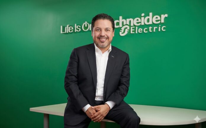 La infraestructura física como pilar estratégico para sostener la acelerada transformación digital impulsada por la IA y la analítica Fotografía de estudio en plano medio que muestra a José Alberto Llavot, directivo de Schneider Electric, posando sentado e inclinado hacia adelante sobre el borde de un escritorio blanco. El ejecutivo, de cabello oscuro y barba ligera, viste un saco de traje negro, camisa blanca de cuello abierto y mantiene las manos entrelazadas al frente con una sonrisa clara hacia la cámara. Detrás de él, un muro liso de color verde brillante exhibe el logotipo institucional de