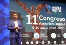 Transformación digital e inteligencia artificial redefinen los negocios en América Digital 2026 Fotografía en plano medio largo que muestra a Lesley Robles, director de América Digital, de pie pronunciando una conferencia magistral sobre un escenario. Viste un traje formal azul de cuadros discretos, camisa blanca de cuello abierto y sostiene un apuntador digital en su mano. Detrás de él, la enorme pantalla de proyección exhibe un fondo con la silueta dorada del Ángel de la Independencia, el título "11° Congreso América Digital - 9-10 Junio 2026 • WTC" y métricas que detallan "+5000 asistentes", "+200 expositores" y "+22 países", acompañadas por íconos de verticales tecnológicas en la base.