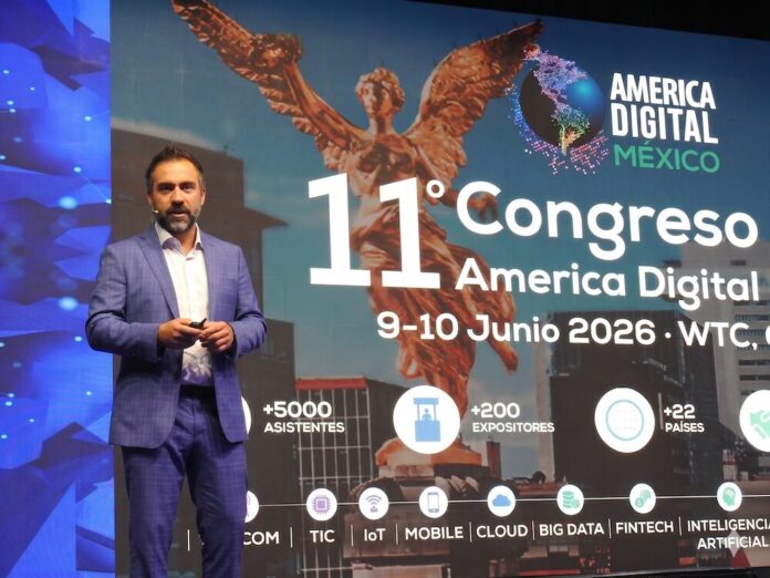 Fotografía en plano medio largo que muestra a Lesley Robles, director de América Digital, de pie pronunciando una conferencia magistral sobre un escenario. Viste un traje formal azul de cuadros discretos, camisa blanca de cuello abierto y sostiene un apuntador digital en su mano. Detrás de él, la enorme pantalla de proyección exhibe un fondo con la silueta dorada del Ángel de la Independencia, el título 