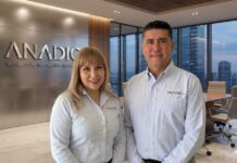 ANADIC: inteligencia artificial para canales TI redefine el negocio tecnológico Fotografía corporativa en plano medio que muestra a Sonia Rodríguez y David Grajales posando sonrientes de pie, uno al lado del otro. Ambos visten camisas formales de manga larga de color azul claro con el logotipo de "ANADIC MX" bordado en el pecho. Se ubican en el interior de una oficina de alta dirección; a la izquierda se aprecia un muro con paneles metálicos que exhibe las letras en relieve de "ANADIC" sobre la leyenda "TECNOLOGÍA • INNOVACIÓN • ESTRATEGIA". Al fondo, a través de un gran ventanal de piso a techo, se observa de manera difuminada una sala de juntas con una mesa de madera y una vista panorámica hacia los rascacielos de la ciudad al atardecer.
