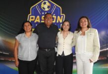 Ingram Micro apuesta por los canales de tecnología con un modelo integral de crecimiento Fotografía grupal de plano entero que captura a cuatro ejecutivos (un hombre y tres mujeres) alineados de pie y sonriendo frente a la cámara. De izquierda a derecha: una especialista con blusa a rayas grises y pantalón negro, el directivo de Ingram Micro con camisa gris de puntos, una ejecutiva con chaleco blanco y pantalón oscuro, y una directiva vestida con un saco texturizado claro y pantalón blanco. Posan en un set corporativo delante de una mampara publicitaria de fondo azul y negro con gráficos deportivos de fútbol.