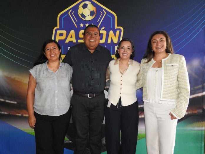 Líderes y especialistas de Ingram Micro formalizan iniciativas de transformación digital Fotografía grupal de plano entero que captura a cuatro ejecutivos (un hombre y tres mujeres) alineados de pie y sonriendo frente a la cámara. De izquierda a derecha: una especialista con blusa a rayas grises y pantalón negro, el directivo de Ingram Micro con camisa gris de puntos, una ejecutiva con chaleco blanco y pantalón oscuro, y una directiva vestida con un saco texturizado claro y pantalón blanco. Posan en un set corporativo delante de una mampara publicitaria de fondo azul y negro con gráficos deportivos de fútbol.