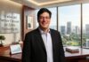 Partner Connect: 10 años redefinen la estrategia de Zebra Technologies Fotografía corporativa en plano medio y de frente de Mauricio López, Vicepresidente de canales para América Latina en Zebra Technologies. El ejecutivo, sonriente y con lentes de armazón delgado, viste un saco negro formal sobre una camisa azul claro de cuello abierto. Se encuentra de pie en una oficina de alta dirección detrás de un escritorio de madera ejecutiva que sostiene una tableta digital con gráficas de tendencias y un fajo de documentos. A la izquierda, un muro texturizado exhibe una placa metálica con la inscripción "10 años del Partner Connect de Zebra Technologies". Al fondo, un gran ventanal de piso a techo ofrece una vista panorámica iluminada de rascacielos y áreas verdes urbanas durante el día.
