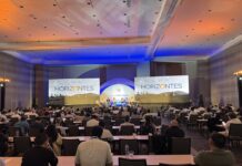 CompuSoluciones Horizontes: el nuevo mapa del canal para crecer con inteligencia artificial, especialización y servicios Fotografía panorámica desde la parte trasera de un gran salón de convenciones lleno de asistentes sentados frente a mesas largas de madera. Al frente se ubica un gran escenario iluminado bajo luces de acento azules y naranjas, donde se desarrolla un panel con ponentes sentados en fila. El escenario cuenta con dos pantallas gigantes en los extremos que muestran la palabra "HORIZONTES" sobre un paisaje montañoso en blanco y negro, flanqueando un arco central blanco con el logotipo circular multicolor de "CompuSoluciones".