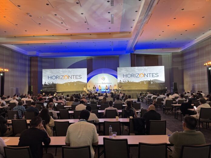 Fotografía panorámica desde la parte trasera de un gran salón de convenciones lleno de asistentes sentados frente a mesas largas de madera. Al frente se ubica un gran escenario iluminado bajo luces de acento azules y naranjas, donde se desarrolla un panel con ponentes sentados en fila. El escenario cuenta con dos pantallas gigantes en los extremos que muestran la palabra 