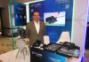 Halo-ProLabs: soluciones para redes de telecomunicaciones en la era digital Fotografía en plano medio de Miguel Cruz, VP de Ventas de Halo-ProLabs, posando solo con una sonrisa ligera y los brazos hacia atrás en un módulo corporativo. Viste un saco formal de tono caqui, camisa blanca de vestir con cuello abierto y anteojos de marco oscuro. En la parte inferior del encuadre se observa la barra de atención con los muestrarios abiertos llenos de transceptores SFP, teniendo detrás una pantalla de televisión digital encendida que despliega una demostración técnica titulada "Installing Prodigy Full Size Converter into Terminal".