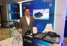 Halo-ProLabs y soluciones para redes de telecomunicaciones en la era digital Fotografía en plano medio de Miguel Cruz, VP de Ventas de Halo-ProLabs, posando solo con una sonrisa ligera y los brazos hacia atrás en un módulo corporativo. Viste un saco formal de tono caqui, camisa blanca de vestir con cuello abierto y anteojos de marco oscuro. En la parte inferior del encuadre se observa la barra de atención con los muestrarios abiertos llenos de transceptores SFP, teniendo detrás una pantalla de televisión digital encendida que despliega una demostración técnica titulada "Installing Prodigy Full Size Converter into Terminal".
