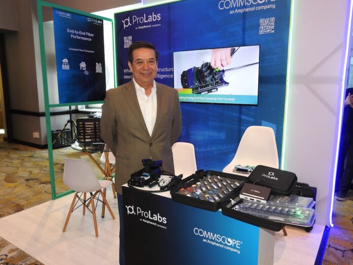 Miguel Cruz delinea la estrategia de Halo-ProLabs para impulsar la eficiencia operativa de los operadores de telecomunicaciones Fotografía en plano medio de Miguel Cruz, VP de Ventas de Halo-ProLabs, posando solo con una sonrisa ligera y los brazos hacia atrás en un módulo corporativo. Viste un saco formal de tono caqui, camisa blanca de vestir con cuello abierto y anteojos de marco oscuro. En la parte inferior del encuadre se observa la barra de atención con los muestrarios abiertos llenos de transceptores SFP, teniendo detrás una pantalla de televisión digital encendida que despliega una demostración técnica titulada