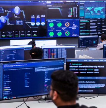 Fortinet presenta FortiSOC, su nueva plataforma SOC unificada impulsada por IA agéntica Fotografía en plano general largo, tomada desde una perspectiva trasera elevada, que muestra el interior de un centro de operaciones de seguridad (SOC) de Fortinet de alta tecnología. En primer plano, la silueta desenfocada de la nuca y el rostro de perfil de un analista frente a su monitor introduce la escena. Múltiples ingenieros y técnicos de seguridad trabajan en sus estaciones individuales equipadas con pantallas de visualización de datos. Al frente, un gran videowall o panel central de pantallas gigantes despliega el panel de control unificado de FortiSOC, mostrando gráficos tridimensionales de flujos de amenazas, mapas globales de ataques en tiempo real, barras de rendimiento operativo y una alerta de brote crítico de ciberseguridad titulada "Microsoft SharePoint Zero-day Attack".
