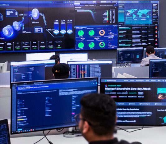 Fortinet presenta FortiSOC, su nueva plataforma SOC unificada impulsada por IA agéntica Fotografía en plano general largo, tomada desde una perspectiva trasera elevada, que muestra el interior de un centro de operaciones de seguridad (SOC) de Fortinet de alta tecnología. En primer plano, la silueta desenfocada de la nuca y el rostro de perfil de un analista frente a su monitor introduce la escena. Múltiples ingenieros y técnicos de seguridad trabajan en sus estaciones individuales equipadas con pantallas de visualización de datos. Al frente, un gran videowall o panel central de pantallas gigantes despliega el panel de control unificado de FortiSOC, mostrando gráficos tridimensionales de flujos de amenazas, mapas globales de ataques en tiempo real, barras de rendimiento operativo y una alerta de brote crítico de ciberseguridad titulada "Microsoft SharePoint Zero-day Attack".
