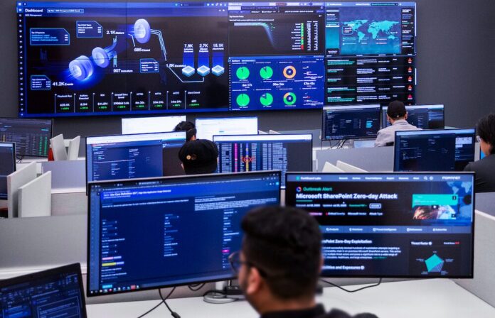 Fotografía en plano general largo, tomada desde una perspectiva trasera elevada, que muestra el interior de un centro de operaciones de seguridad (SOC) de Fortinet de alta tecnología. En primer plano, la silueta desenfocada de la nuca y el rostro de perfil de un analista frente a su monitor introduce la escena. Múltiples ingenieros y técnicos de seguridad trabajan en sus estaciones individuales equipadas con pantallas de visualización de datos. Al frente, un gran videowall o panel central de pantallas gigantes despliega el panel de control unificado de FortiSOC, mostrando gráficos tridimensionales de flujos de amenazas, mapas globales de ataques en tiempo real, barras de rendimiento operativo y una alerta de brote crítico de ciberseguridad titulada 