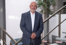 Oscar Martínez, de Kingston Technology: “Hoy, la resiliencia digital es la base de la continuidad operativa” Fotografía en plano medio y de frente de Óscar Martínez, Regional Director de Kingston Technology, posando sonriente en un entorno de oficina iluminado con luz natural de día. El directivo, calvo y con barba corta canosa, viste un saco formal gris texturizado a cuadros sobre una camisa blanca de cuello abierto. Se encuentra de pie sosteniendo sus manos entrelazadas al frente, con su cuerpo apoyado ligeramente sobre el barandal metálico de una escalera industrial de peldaños gris claro. Al fondo se aprecia de forma difuminada un patio interior con un árbol decorativo.