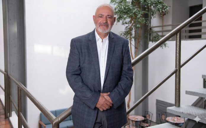 Fotografía en plano medio y de frente de Óscar Martínez, Regional Director de Kingston Technology, posando sonriente en un entorno de oficina iluminado con luz natural de día. El directivo, calvo y con barba corta canosa, viste un saco formal gris texturizado a cuadros sobre una camisa blanca de cuello abierto. Se encuentra de pie sosteniendo sus manos entrelazadas al frente, con su cuerpo apoyado ligeramente sobre el barandal metálico de una escalera industrial de peldaños gris claro. Al fondo se aprecia de forma difuminada un patio interior con un árbol decorativo.