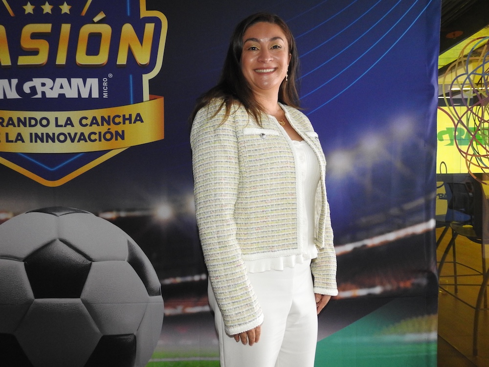 Fotografía en plano medio de Paula Contreras, IMFS Regional Lead Latam, posando sonriente en un entorno corporativo de tres cuartos hacia la izquierda. Viste una chaqueta corta texturizada en tonos claros sobre una blusa blanca y pantalón formal blanco. A su izquierda, en el fondo, se observa un cartel publicitario azul marino con el escudo amarillo de "PASIÓN INGRAM" y un balón de fútbol en primer plano.