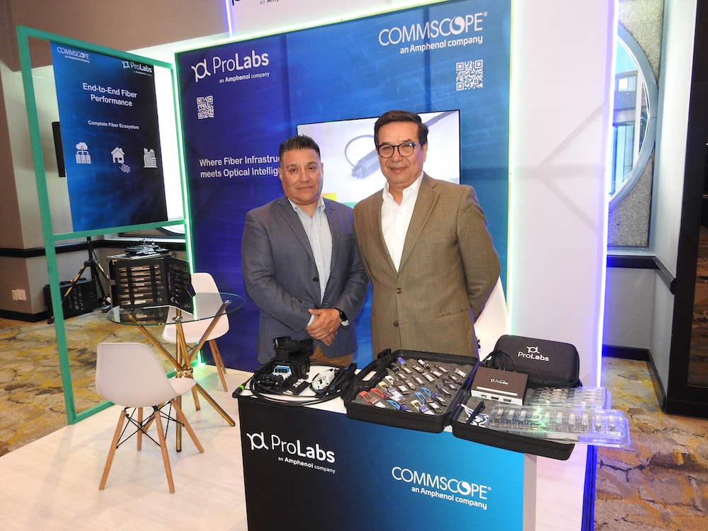 soluciones para redes de telecomunicaciones: Fotografía en plano medio que muestra a dos ejecutivos de pie y sonrientes dentro de un stand de exhibición comercial de ProLabs y CommScope. El profesional de la izquierda viste un saco gris azulado, mientras que el de la derecha usa un saco color marrón claro y anteojos. En frente de ellos, sobre un mostrador negro con el logotipo de ambas marcas, se aprueban maletines abiertos que exhiben una variedad de módulos transceptores ópticos SFP multimarca y equipos de conectividad. El fondo cuenta con paneles azules iluminados por tiras LED verdes que muestran los títulos "ProLabs" y "COMMSCOPE an Amphenol company" junto a códigos QR.