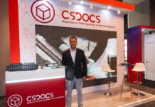 CS Docs lleva la digitalización documental empresarial más allá del papel Fotografía en plano general medio que muestra a Rafael Cotera Reynoso, gerente comercial de CS Docs, posando de frente con una postura formal en el stand corporativo de la empresa. El ejecutivo, de tez morena y cabello corto oscuro, viste un saco de traje negro sobre una camisa de vestir de rayas finas y pantalón claro. A su izquierda, sobre un mostrador rojo brillante con el logotipo blanco de "CS Docs Mayorista en Ciberseguridad e Hiperconvergencia", se exhiben varios equipos físicos de red y servidores de almacenamiento. Al fondo resalta una gran pantalla digital y dos bancos altos de color blanco junto a una mesa redonda de cristal.