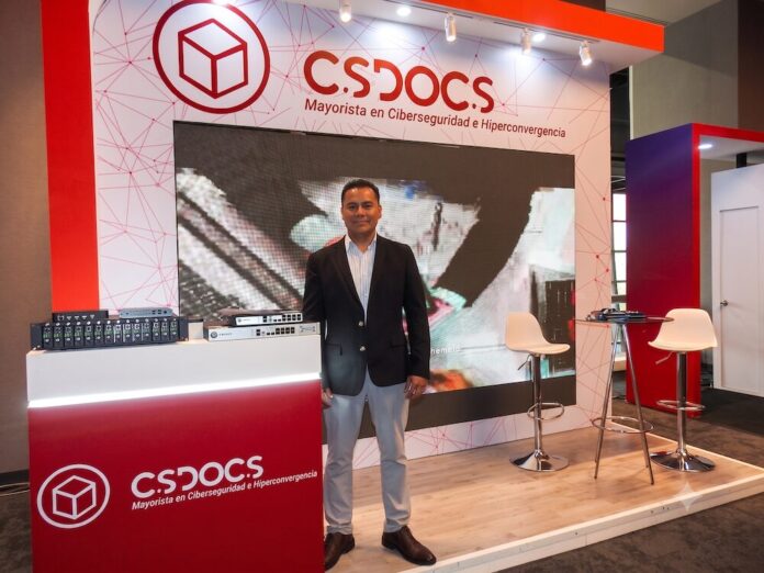 Fotografía en plano general medio que muestra a Rafael Cotera Reynoso, gerente comercial de CS Docs, posando de frente con una postura formal en el stand corporativo de la empresa. El ejecutivo, de tez morena y cabello corto oscuro, viste un saco de traje negro sobre una camisa de vestir de rayas finas y pantalón claro. A su izquierda, sobre un mostrador rojo brillante con el logotipo blanco de 