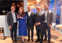 Red Hat reconoce innovación del IMSS: una plataforma capaz de procesar miles de millones de transacciones diarias Fotografía grupal en plano entero de cinco directivos y especialistas de Red Hat y el IMSS posando de pie alineados en una sala corporativa. Al extremo izquierdo figura Javier Cordero y a su lado una ejecutiva con vestido azul rey sostiene un galardón transparente de reconocimiento. Los acompañan tres ejecutivos con trajes oscuros y sacos formales. Al fondo se aprecian ventanales con vista urbana al atardecer y un par de taburetes altos.