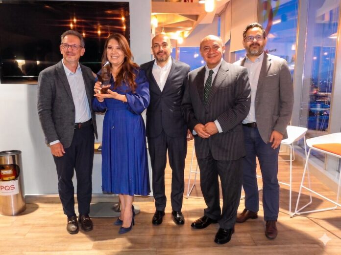 Red Hat entrega reconocimiento oficial al IMSS por consolidar una estrategia integral de evolución tecnológica Fotografía grupal en plano entero de cinco directivos y especialistas de Red Hat y el IMSS posando de pie alineados en una sala corporativa. Al extremo izquierdo figura Javier Cordero y a su lado una ejecutiva con vestido azul rey sostiene un galardón transparente de reconocimiento. Los acompañan tres ejecutivos con trajes oscuros y sacos formales. Al fondo se aprecian ventanales con vista urbana al atardecer y un par de taburetes altos.