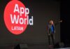F5 AppWorld LATAM 2026: la IA pasa de la experimentación a la operación empresarial Fotografía en plano medio largo que muestra a Roberto Ricossa, vicepresidente de F5 para Latinoamérica, de pie en un escenario oscuro durante su presentación inaugural. El ejecutivo viste un saco gris, playera azul brillante, pantalones oscuros y tenis con detalles rojos, y aparece con el brazo izquierdo extendido señalando con el dedo índice hacia el frente. A su derecha se aprecia una enorme pantalla LED negra que despliega un gran círculo rojo con el logotipo del evento en letras blancas: "App World LATAM". Sobre la tarima descansan dos monitores de piso negros en primer plano.
