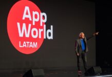 F5 AppWorld LATAM 2026: la IA pasa de la experimentación a la operación empresarial Fotografía en plano medio largo que muestra a Roberto Ricossa, vicepresidente de F5 para Latinoamérica, de pie en un escenario oscuro durante su presentación inaugural. El ejecutivo viste un saco gris, playera azul brillante, pantalones oscuros y tenis con detalles rojos, y aparece con el brazo izquierdo extendido señalando con el dedo índice hacia el frente. A su derecha se aprecia una enorme pantalla LED negra que despliega un gran círculo rojo con el logotipo del evento en letras blancas: "App World LATAM". Sobre la tarima descansan dos monitores de piso negros en primer plano.