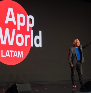 F5 AppWorld LATAM 2026: la IA pasa de la experimentación a la operación empresarial Fotografía en plano medio largo que muestra a Roberto Ricossa, vicepresidente de F5 para Latinoamérica, de pie en un escenario oscuro durante su presentación inaugural. El ejecutivo viste un saco gris, playera azul brillante, pantalones oscuros y tenis con detalles rojos, y aparece con el brazo izquierdo extendido señalando con el dedo índice hacia el frente. A su derecha se aprecia una enorme pantalla LED negra que despliega un gran círculo rojo con el logotipo del evento en letras blancas: "App World LATAM". Sobre la tarima descansan dos monitores de piso negros en primer plano.