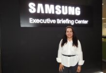 El Showroom de Samsung muestra cómo las soluciones profesionales están transformando el negocio de los canales Retrato corporativo en plano medio de Ana Lira, Directora B2B de Consumer Electronics en Samsung Electronics, posando sonriente con los brazos a los costados. Viste una blusa formal blanca de manga larga con un cinturón delgado negro y pantalón oscuro. Se ubica en el interior de un showroom frente a una pared negra mate que exhibe en la parte superior el letrero corporativo iluminado en letras blancas tridimensionales "SAMSUNG Executive Briefing Center".