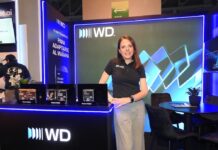 Western Digital: almacenamiento empresarial interno seguro y escalable Fotografía de tres cuartos de Sandra Mendoza, gerente de Mercadotecnia para Latinoamérica en Western Digital, posando de pie con una sonrisa frente a un stand corporativo. Viste una playera polo negra con el logotipo de WD y pantalones de vestir color gris claro. A su izquierda, sobre un mostrador negro con luz LED azul perimetral, se encuentran en exhibición tres discos duros Ultrastar. A su espalda, un gran panel vertical muestra el mensaje "ALMACENAMIENTO PARA ADAPTARSE AL MAÑANA", acompañado de una mesa redonda negra con sillas de terciopelo en el lado derecho del encuadre.