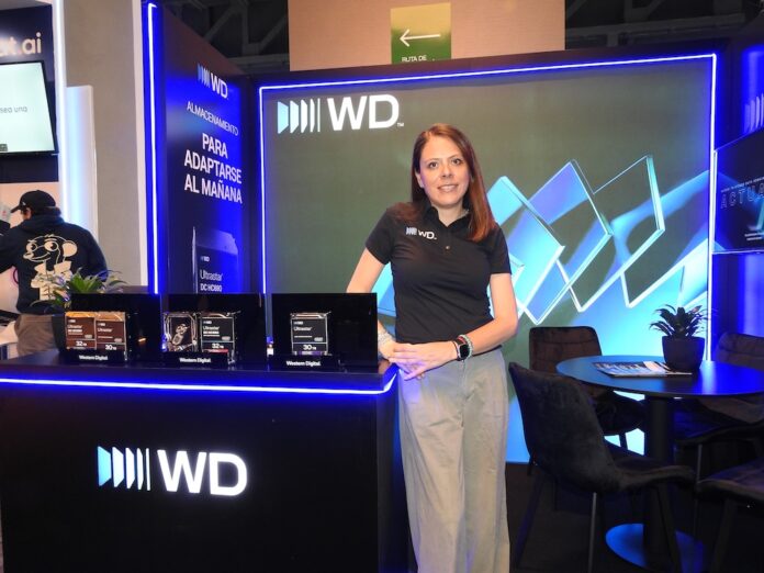Fotografía de tres cuartos de Sandra Mendoza, gerente de Mercadotecnia para Latinoamérica en Western Digital, posando de pie con una sonrisa frente a un stand corporativo. Viste una playera polo negra con el logotipo de WD y pantalones de vestir color gris claro. A su izquierda, sobre un mostrador negro con luz LED azul perimetral, se encuentran en exhibición tres discos duros Ultrastar. A su espalda, un gran panel vertical muestra el mensaje 
