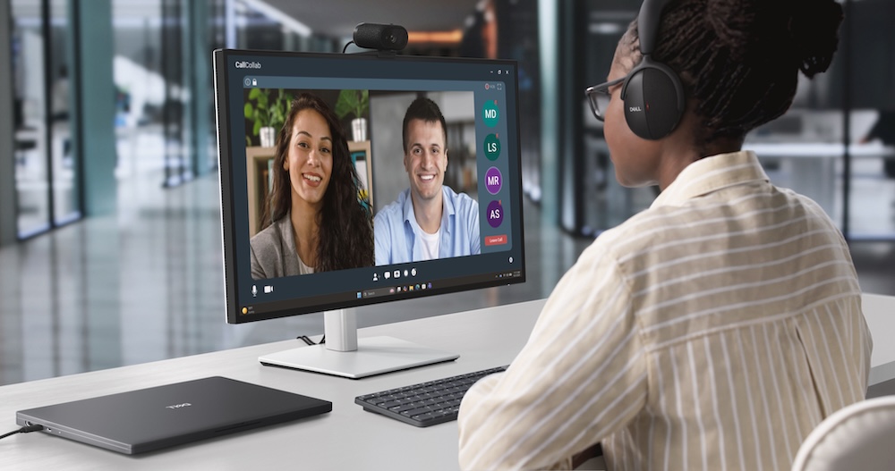 Fotografía de ambiente en plano medio que muestra una computadora todo en uno en un escritorio de diseño minimalista con la cámara web Dell Pro 7 4K instalada en la parte superior. En la pantalla del monitor se observa una interfaz de videollamada activa, mientras que el fondo del espacio de trabajo luce ordenado con una lámpara LED de escritorio y paneles divisorios acústicos de color gris claro.