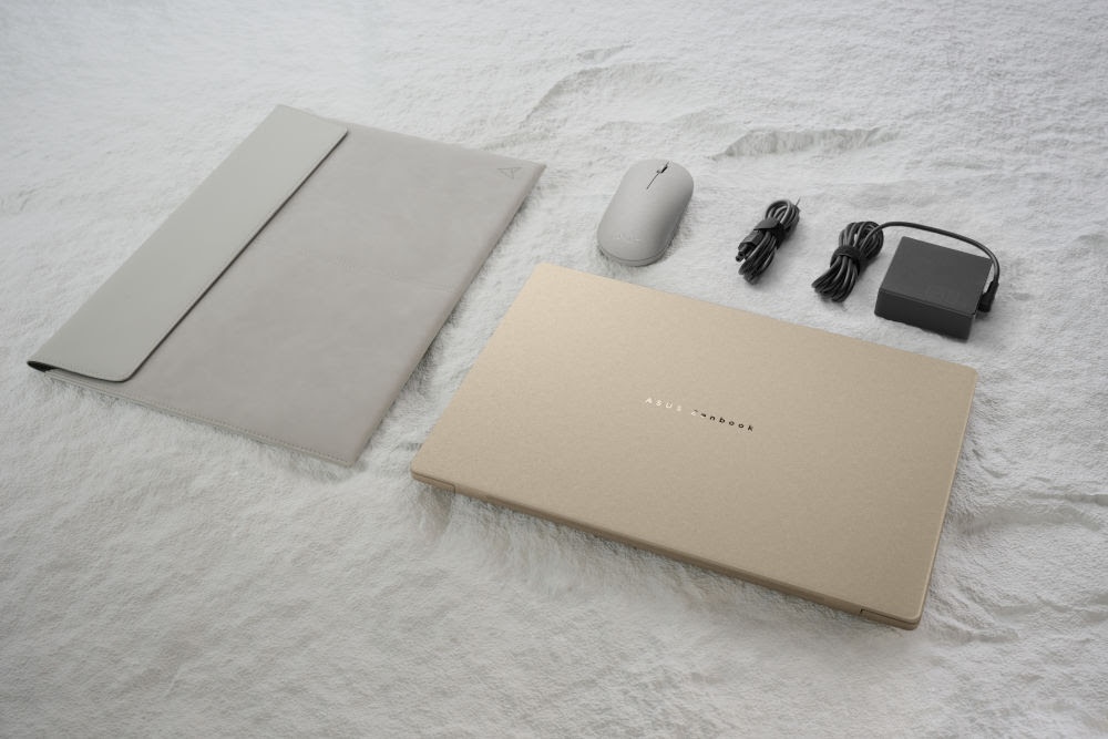 Fotografía cenital de una laptop ASUS Zenbook cerrada en color dorado mate. A su alrededor se muestran sus accesorios organizados: una funda tipo sobre en color gris claro, un mouse inalámbrico del mismo tono y un adaptador de corriente negro con sus cables recogidos. Todos los elementos reposan sobre una superficie blanca con textura rugosa.