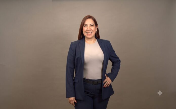 Fotografía de medio cuerpo de una mujer sonriente de cabello castaño rojizo, vistiendo un traje sastre azul marino y blusa color arena, posando con la mano izquierda en la cadera frente a un fondo gris neutro.