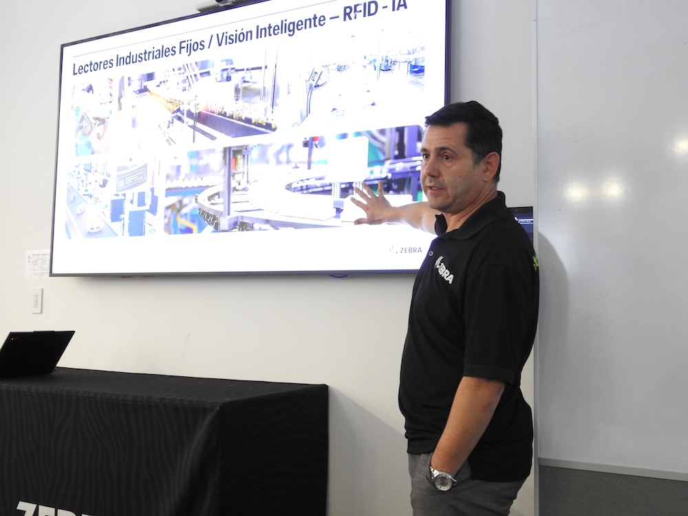Fotografía en plano medio largo que captura a Alfonso Caraveo impartiendo una conferencia técnica en un salón de capacitación. El directivo de Zebra Technologies, vestido con su playera tipo polo negra corporativa, se ubica de perfil extendiendo su brazo izquierdo para señalar una gran pantalla digital empotrada en el muro blanco. La diapositiva proyectada lleva por título "Lectores Industriales Fijos / Visión Inteligente – RFID - IA" y muestra cuatro imágenes de ejemplo de brazos robóticos y bandas transportadoras en plantas de manufactura automatizadas, situándose a la derecha un pintarrón blanco.